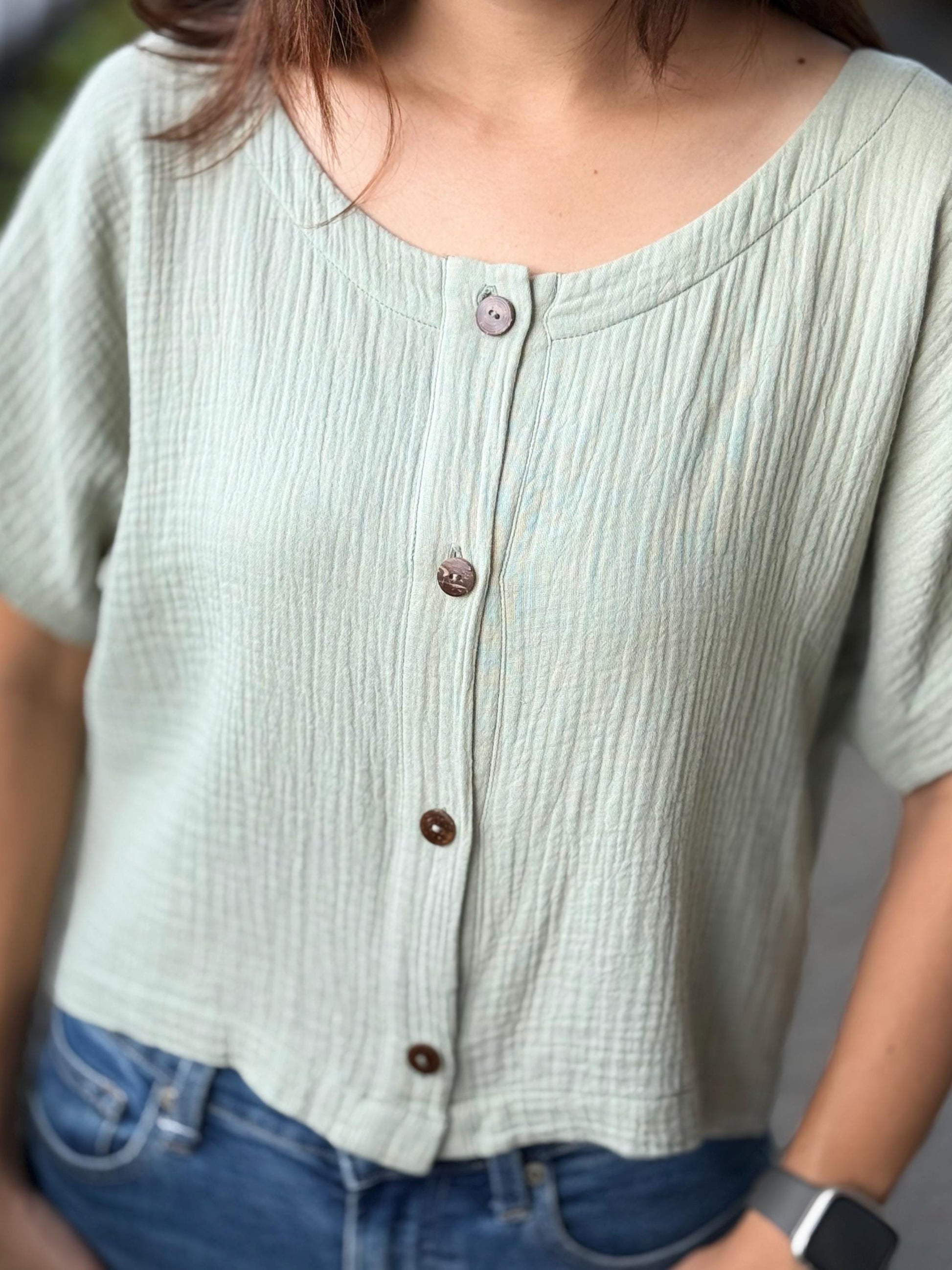 May include: A light sage green cropped button-down top. The top is made of a textured cotton gauze fabric and has short sleeves.  The buttons are dark brown wood. The top has a relaxed, slightly oversized fit.