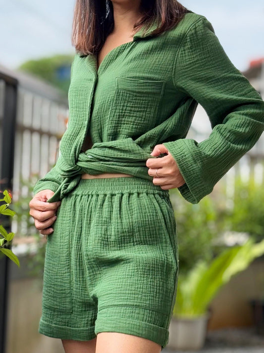 May include: A green cotton short set. The set includes a collared button-up shirt and shorts. The shirt is tied at the waist. The shorts have an elastic waistband. The fabric has a textured appearance.