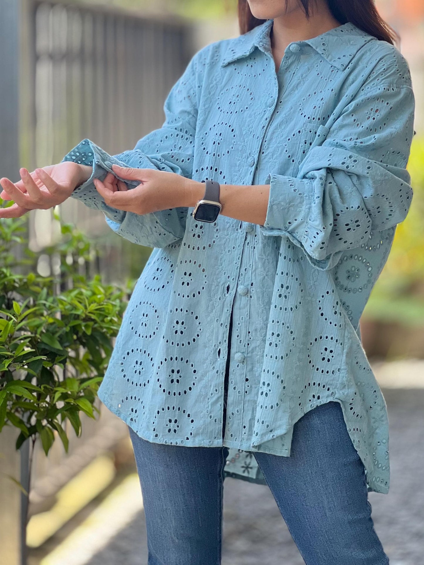 May include: A light blue, long-sleeved, button-up shirt with a delicate eyelet pattern. The shirt has a relaxed fit and a slightly longer hem in the back.